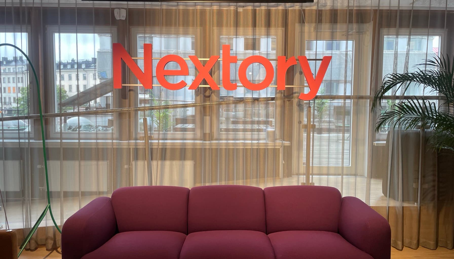The Nextory wordmark in coral lettering across the windows of the company's Stockholm office, overlooking the city's rooftops, with a burgundy sofa below